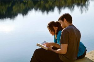 Photo of couple reading Bible beside lake - including God in marriage